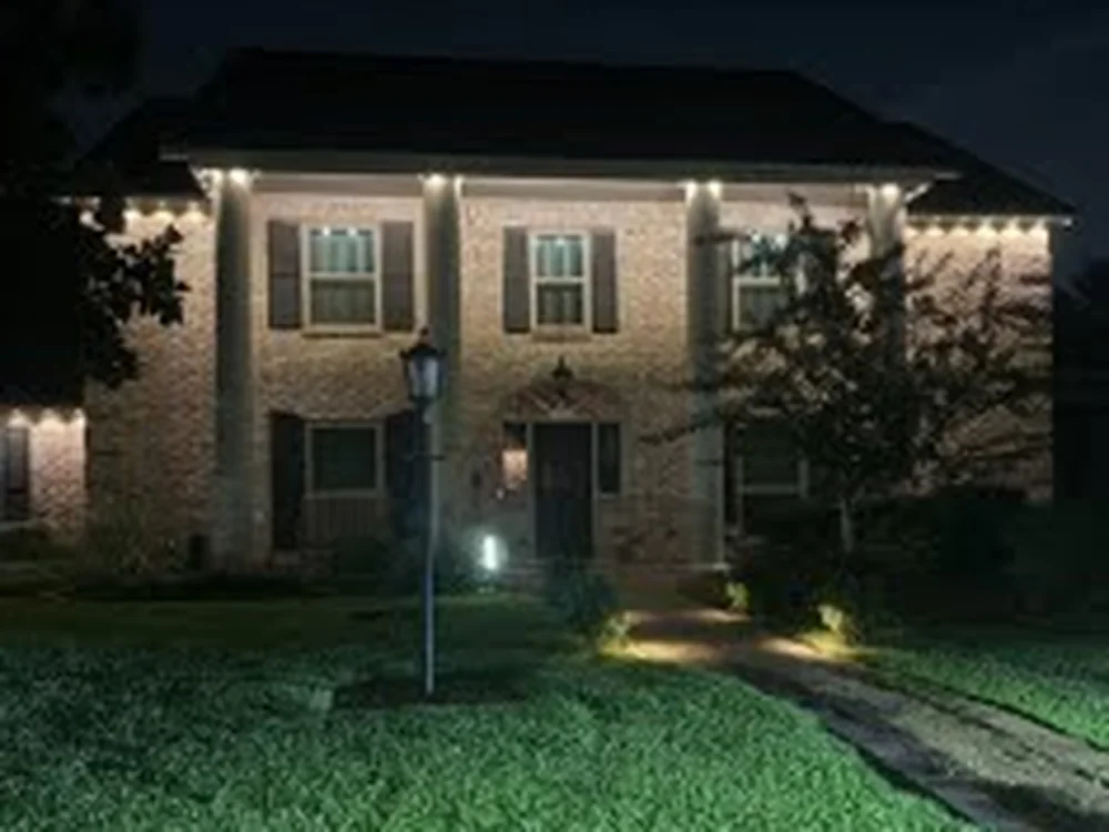 Real Skyclear install — two-story brick home with warm-white permanent lighting outlining the eaves, entry, and a softly lit lawn at night